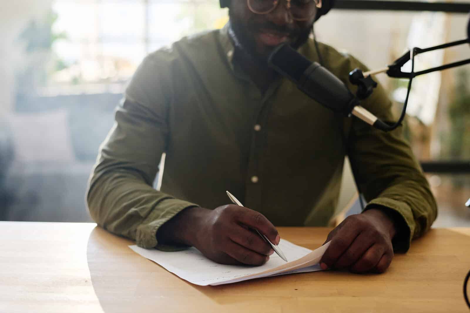 Close-up of young black man pointing at paper with questions for interview and voicing one of them while talking in microphone